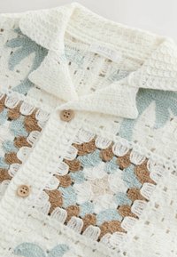 Biały sweterek na szydełku z niebieskimi i brązowymi kwiatowymi wzorami, drewnianymi guzikami oraz kołnierzem złożonym, oznaczony metką "NEXT Made in China".