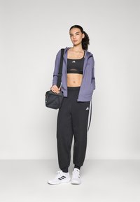 Idrettsantrekk bestående av en lilla jakke med glidelås, svart kort sports-bh, svarte joggebukser med hvite striper og hvite sneakers.
