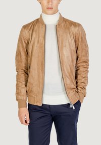 Chaqueta bomber de ante color beige con cierre frontal, puños de canalé y bolsillos laterales, superpuesta a un cuello de tortuga blanco y pantalones azul marino.