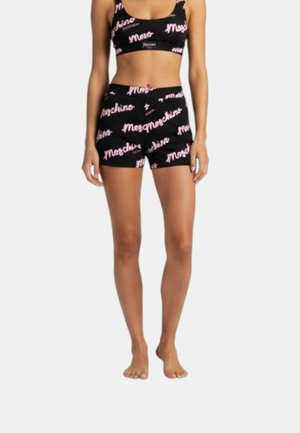 Donna che indossa un completo nero a due pezzi con stampa del logo "Moschino" rosa, composto da crop top e pantaloncini a vita alta, in piedi a piedi nudi.