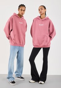 To personer iført matchende overdimensjonerte rosa "OFF LIMITS" hettegensere, en med lyseblå jeans og svarte joggesko, den andre med svarte flarebukser og joggesko.