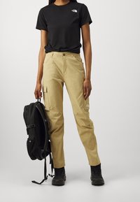 Beige cargobyxor med sidofickor, i kombination med en svart t-shirt med kort ärm. En svart ryggsäck hålls i ena handen. Svarta stövlar fulländar looken.