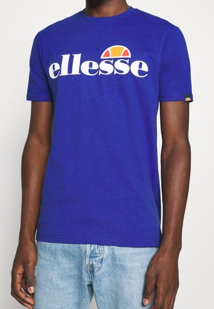 Mann trägt ein leuchtend blaues T-Shirt mit Rundhalsausschnitt, weißem "ellesse"-Logo und orangefarbenem Halbrund-Design, kombiniert mit hellblauen Jeans.