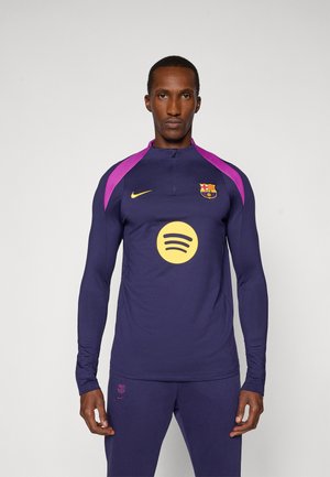 Maglietta sportiva a maniche lunghe di colore viola scuro con dettagli rosa sulle spalle, caratterizzata da un logo Spotify giallo e un piccolo emblema del club. Design aderente.