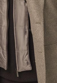 Cappotto a strati con uno strato esterno in lana marrone texturizzata e una fodera interna liscia e imbottita beige, con bottoni e cerniere visibili.
