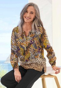 Langärmlige Bluse mit mehrfarbigem Blumen- und Tierdruck, Knopfleiste, weichem Stoff und abgerundetem Saum, kombiniert mit schwarzen Hosen.