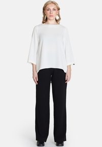 Weißes, lockeres Oberteil mit drei Viertelärmeln, kombiniert mit schwarzen, weiten Hosen. Das Modell präsentiert ein schlichtes, elegantes Outfit.