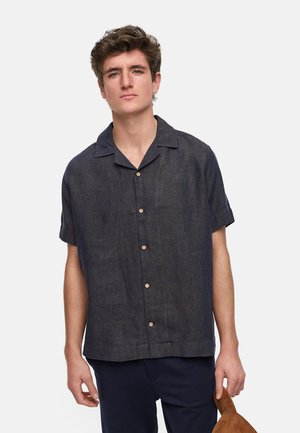 Camisa de lino azul oscuro de manga corta con collar tipo punto, botones horizontales y un corte relajado, que presenta una textura sutil y líneas limpias.
