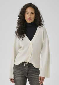 Cream cardigan with a V-neck, button front, ribbed cuffs, and a relaxed fit, worn over a black turtleneck and paired with dark jeans.
