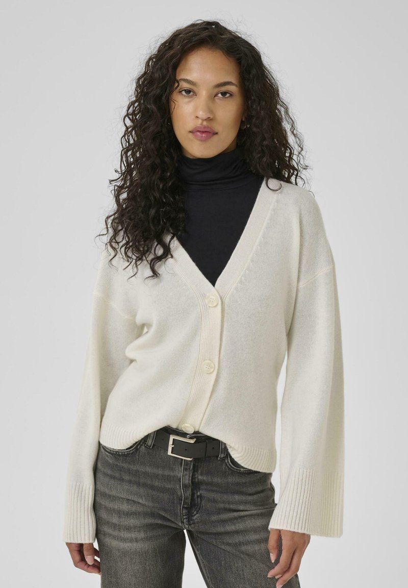 Cream cardigan with a V-neck, button front, ribbed cuffs, and a relaxed fit, worn over a black turtleneck and paired with dark jeans.