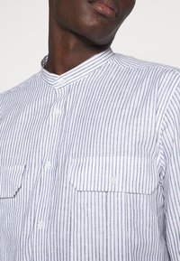 Camicia bianca con strisce verticali blu, colletto a bandiera e due tasche sul petto. Realizzata in un tessuto leggero, con chiusura a bottoni.