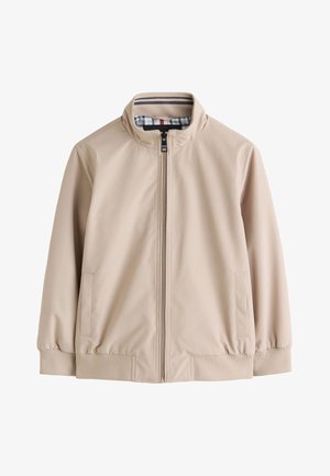 Dreng stående iført beige lynlåsjakke over hvid poloshirt, sorte bukser og hvide sneakers mod ensfarvet beige baggrund.