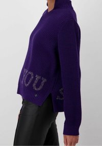 Maglione viola lavorato a maglia con collo alto e spacchi laterali. Presenta una scritta lucida e metallica sulla parte inferiore anteriore che recita "YOU."