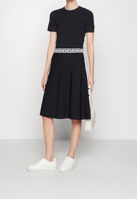 Abito nero a maniche corte con top aderente e gonna svasata, caratterizzato da una cintura bianca con logo. Abbinato a sneaker bianche e una borsa.