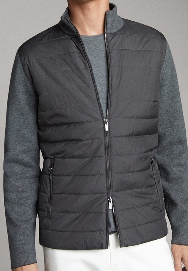 Giacca imbottita con un davanti grigio trapuntato e maniche in maglia grigio scuro. Caratterizzata da un collo alto, chiusura con zip e tasche laterali.