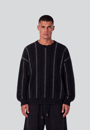 Mann mit kurzen Haaren, der einen schwarzen Pullover mit vertikalen grauen Streifen und schwarze Hose trägt, steht vor einem einfarbigen hellgrauen Hintergrund.