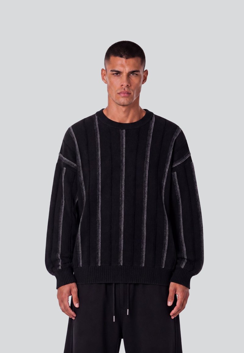 Homme aux cheveux courts portant un pull noir à rayures verticales grises et un pantalon noir, debout devant un fond uni gris clair.