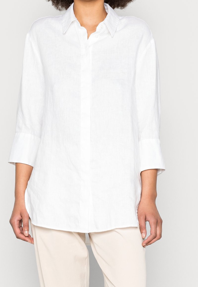 Chemise en lin blanc avec un col à boutons, manches longues retroussées jusqu'aux coudes, et une silhouette droite, présentant une texture subtile.