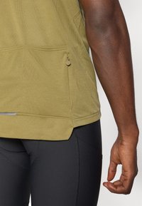 Olivgrünes, mesh Athletikoberteil mit einer seitlichen Reißverschlusstasche, kombiniert mit schwarzen, eng anliegenden Leggings. Verfügt über einen abgerundeten Saum und reflektierende Akzente.