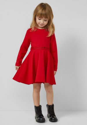 Czerwona sukienka na długi rękaw z rozkloszowaną spódnicą i fakturowanym czarnym paskiem. Do tego błyszczące czarne botki na sznurowadła, stojące na neutralnym tle.