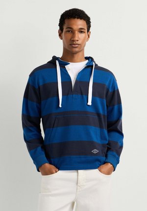 Junger Mann trägt einen blau-navy gestreiften Hoodie mit weißen Kordeln und hellen Hosen, steht vor einem einfarbigen hellen Hintergrund.