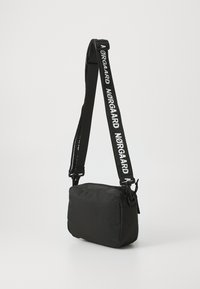 Svart rektangulær crossbody-veske laget av myk tekstil. Justerbar stropp med hvit "NØRGAARD"-merking. Glidelås på toppen.