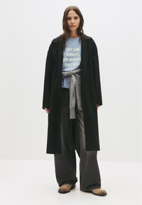 Čierny dlhý kabát, svetlomodrá košeľa s textom a šedý sveter previazaný okolo pása. Tmavé široké nohavice a béžová obuv. Minimalistický dizajn.