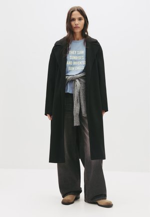 Schwarzer langer Mantel, hellblaue Bluse mit Text und grauer Pullover um die Taille gebunden. Dunkle weit geschnittene Hose und beiges Schuhwerk. Minimalistisches Design.