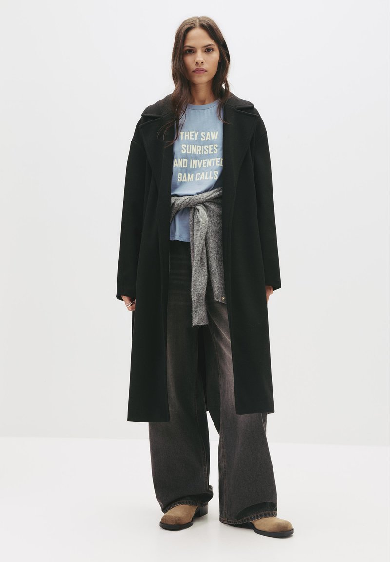 Čierny dlhý kabát, svetlomodrá košeľa s textom a šedý sveter previazaný okolo pása. Tmavé široké nohavice a béžová obuv. Minimalistický dizajn.