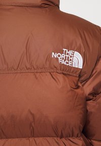 Veste matelassée couleur rouille avec logo "The North Face" brodé en blanc sur le côté droit du haut du dos.