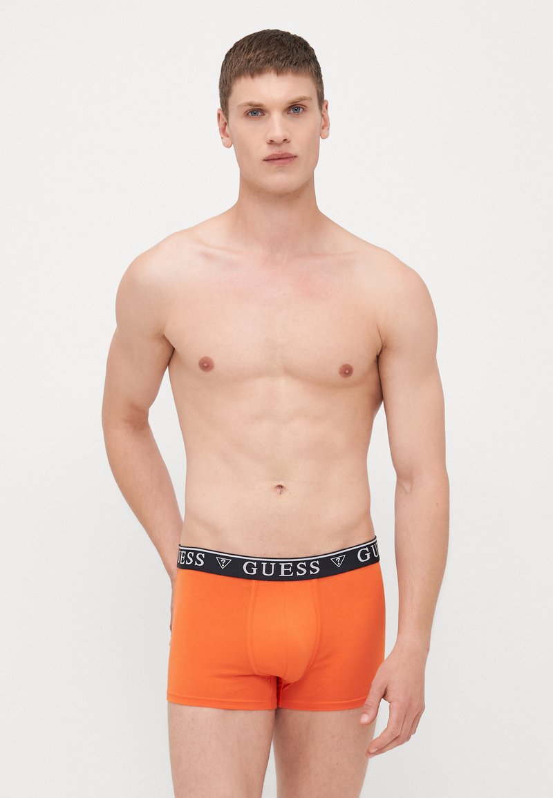 Boxers oranges avec une ceinture noire portant le logo "GUESS". Le tissu semble lisse et extensible, avec un design ajusté.