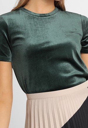 Nainen, joka pukeutunut tummanvihreään samettiseen lyhythihaiseen toppiin, joka on tungettu korkeavyötäröisen, beigen ja mustan laskostetun hameen sisään.