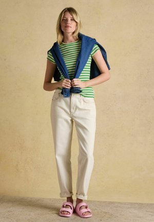 Mladá žena stojaca so založenými rukami, oblečená v zelenom pruhovanom tričku, bielych nohaviciach, modrej bunde prehodenou cez plecia a ružových sandáloch.