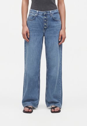 Person trägt hellblaue Jeans mit weitem Bein und sichtbarem Knopfverschluss sowie schwarze Sandalen mit offenen Zehen und steht vor einem einfarbigen Hintergrund.