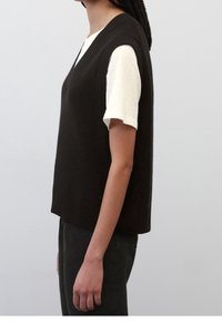 Widok boczny osoby noszącej czarny sweter bez rękawów na białej koszuli z krótkim rękawem, w zestawieniu z czarnymi spodniami na jednolitym tle.