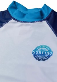 Långärmad surftröja i vitt, med blå detaljer på kragen och ärmarna. Har en cirkulär blå logotyp med texten "Surfing".