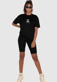 Sort bomulds-t-shirt med korte ærmer og et hvidt grafisk logo på brystet, parret med sorte cykelshorts og beige sneakers.