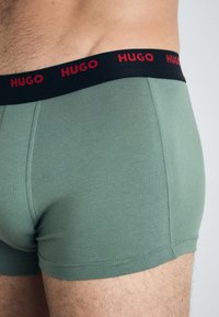 Grüne Baumwoll-Boxershorts mit schwarzem Bund, der rote "HUGO"-Schriftzüge aufweist. Glatte Textur mit flachen Nähten, körperbetonter Schnitt.