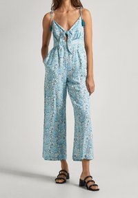 Modrý overal s vzorem, s vázáním vpředu, nastavitelnými ramínky a širokými nohavicemi, zdobený květinovými motivy a měkkou texturou látky. Černé sandály na podpatku.