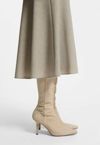Botas beige hasta la rodilla con acabado en suave ante, que presentan punteras puntiagudas y tacones de aguja. Combinadas con una falda fluida de color gris claro.