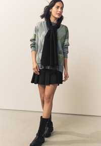 Hyggelig grå sweater med grønt diamantmønster, sort frynset tørklæde, sort plisseret nederdel og sort knæhøje støvler med spænde-detaljer.