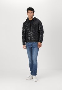 Giubbotto puffer nero con finitura lucida, chiusura con zip, cappuccio e tasche laterali. Abbinato a jeans blu e sneaker bianche su uno sfondo neutro.