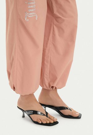 Piedi nudi che indossano sandali neri con tacco e cinturini con strass "Juicy", abbinati a pantaloni rosa larghi raccolti alle caviglie.