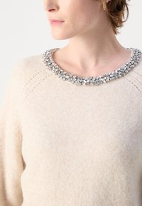 Pull beige avec une finition texturée, présentant un décolleté orné de pierres précieuses claires de différentes formes.