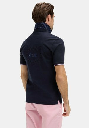 Homme portant un polo bleu marine avec le col relevé dévoilant un tissu intérieur à motifs et un short rose, vu de dos.