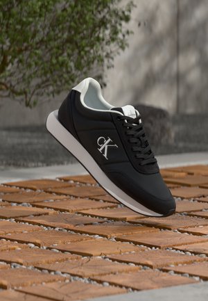 Baskets Calvin Klein noir et blanc flottant au-dessus d'un sol en bois et gravier avec des plantes et un mur flous en arrière-plan.