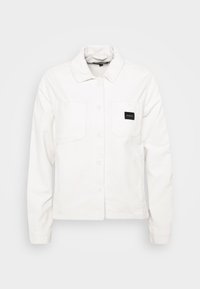 Vit kordjacka med knäppning framtill, två bröstfickor och en svart etikett på vänster ficka. Har en klassisk kragdesign.