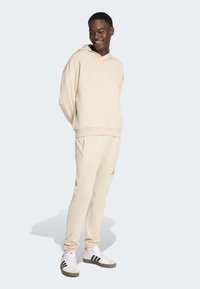 Mladý muž se usmívá, má na sobě béžovou mikinu a tepláky s bílými teniskami Adidas, stojí před světle šedým pozadím.