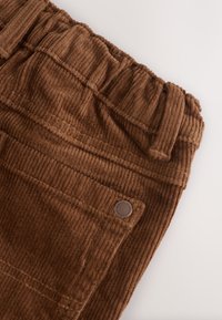 Pantalon en velours côtelé marron avec taille élastique, passants pour ceinture, poche pour les doigts et détails de bouton métallique sur fond clair.