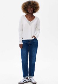 Femme avec des cheveux bouclés portant un pull blanc à col en V, un jean bleu retroussé, et des baskets blanches, se tenant avec une main dans la poche.
